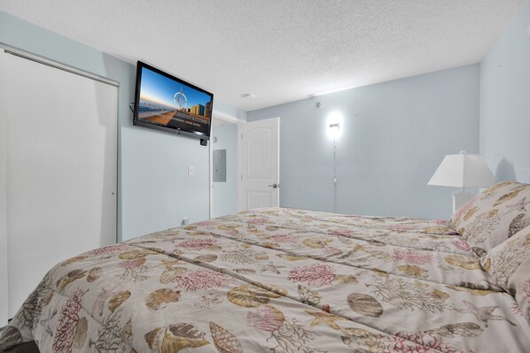 Un dormitorio con una cama, un televisor instalado en la pared, una puerta y una lámpara de pared.