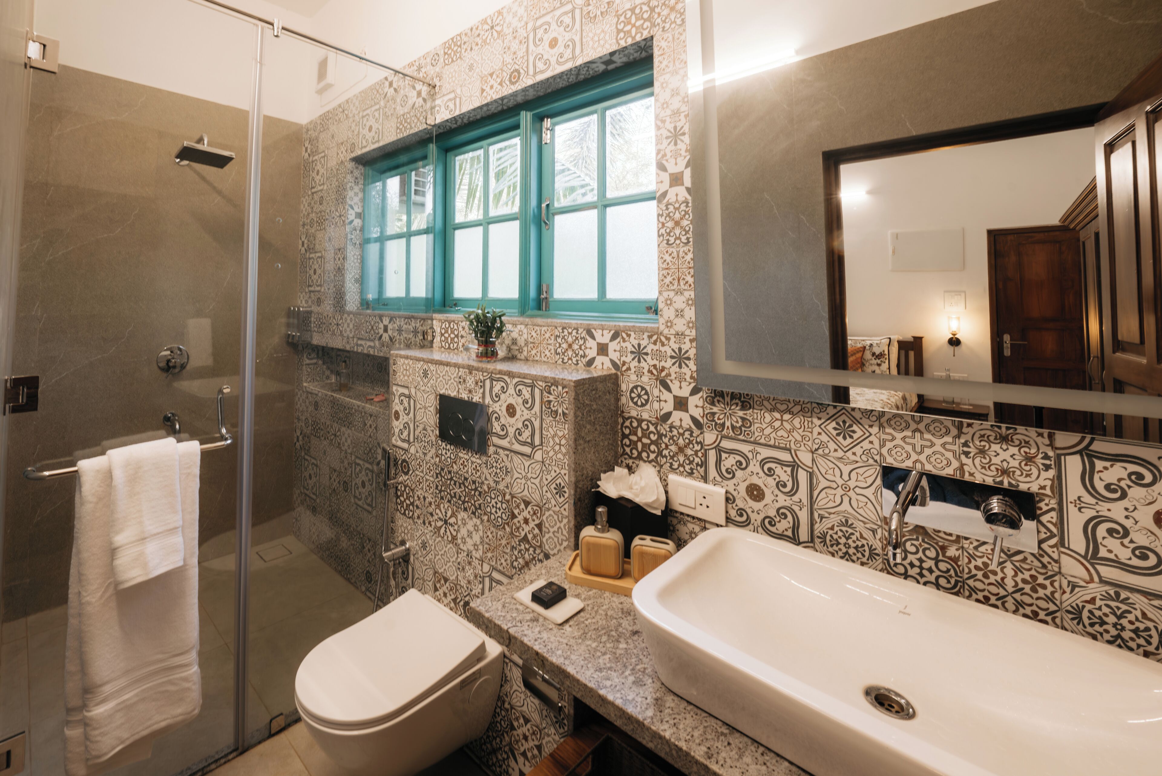 A bathroom with a glass shower enclosure, a toilet, a sink, and a window with a teal frame.