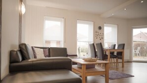 A modern living room with a sofa, a dining table, and chairs. There are large windows and a view of the outdoors. - Beautiful vacation home "Nele"! Perfect for the family (Geeste)