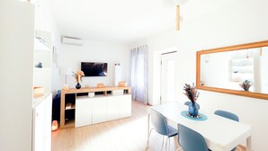A modern, well-lit living room with a TV, wooden furniture, and a dining area with chairs and a table.