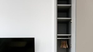 Una habitación con televisión montada en la pared, un banco de madera y un armario con ropa colgada.