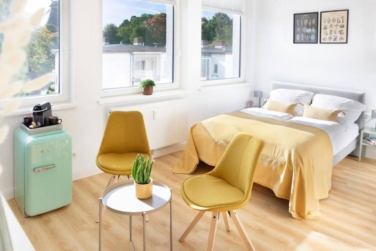 Un dormitorio moderno con una cama, dos sillas, una mesita, un refrigerador antiguo y un ventanal con vistas a edificios y árboles.