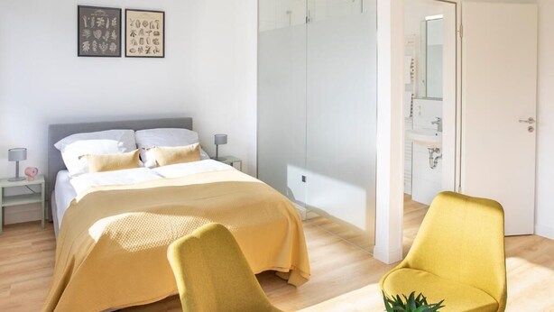 Un dormitorio moderno con una cama, un sillón amarillo, un box de ducha de vidrio y una puerta blanca que da al baño.