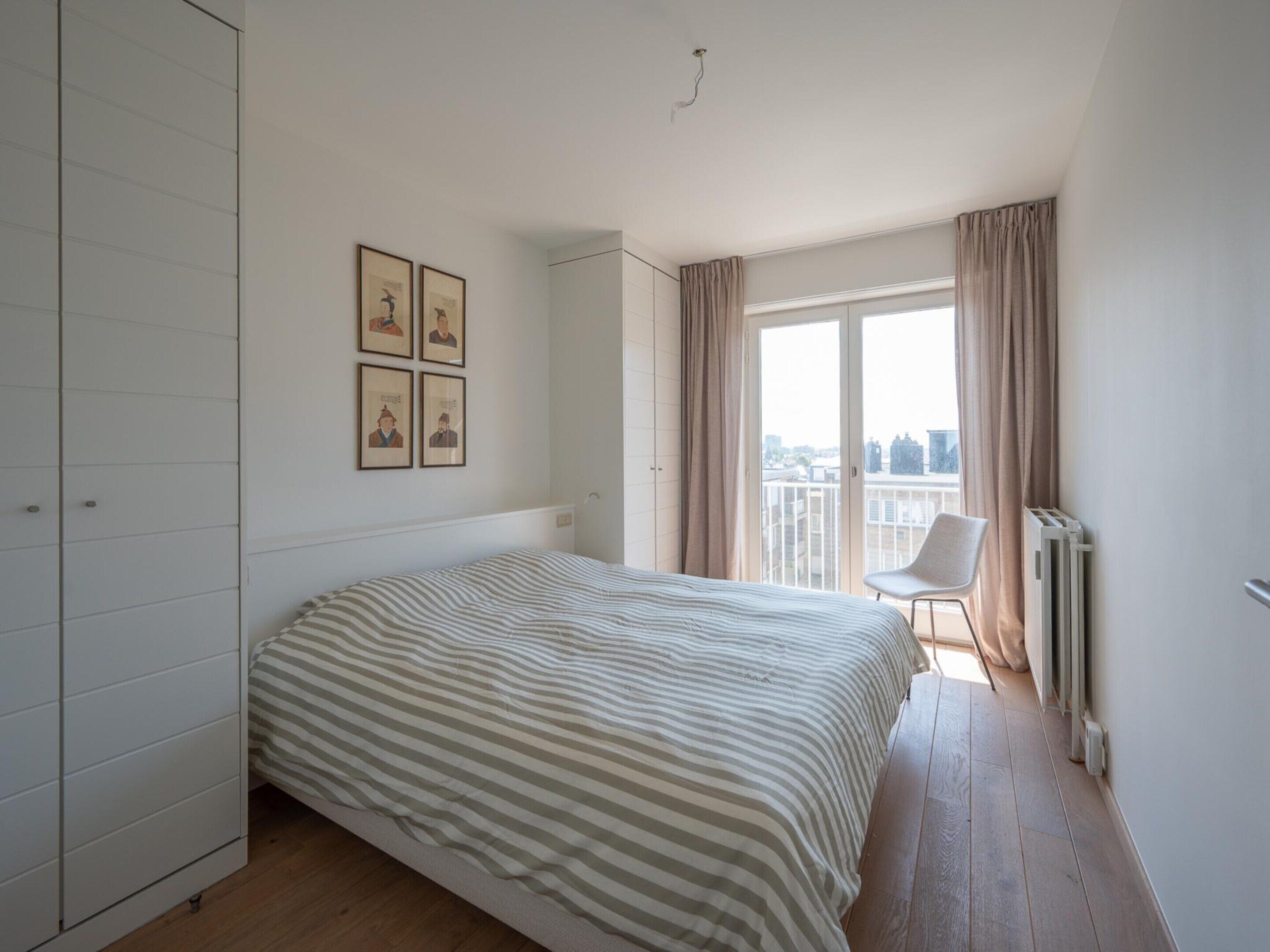 Un dormitorio con cama, una silla, un radiador y vistas a la ciudad a través de una ventana.