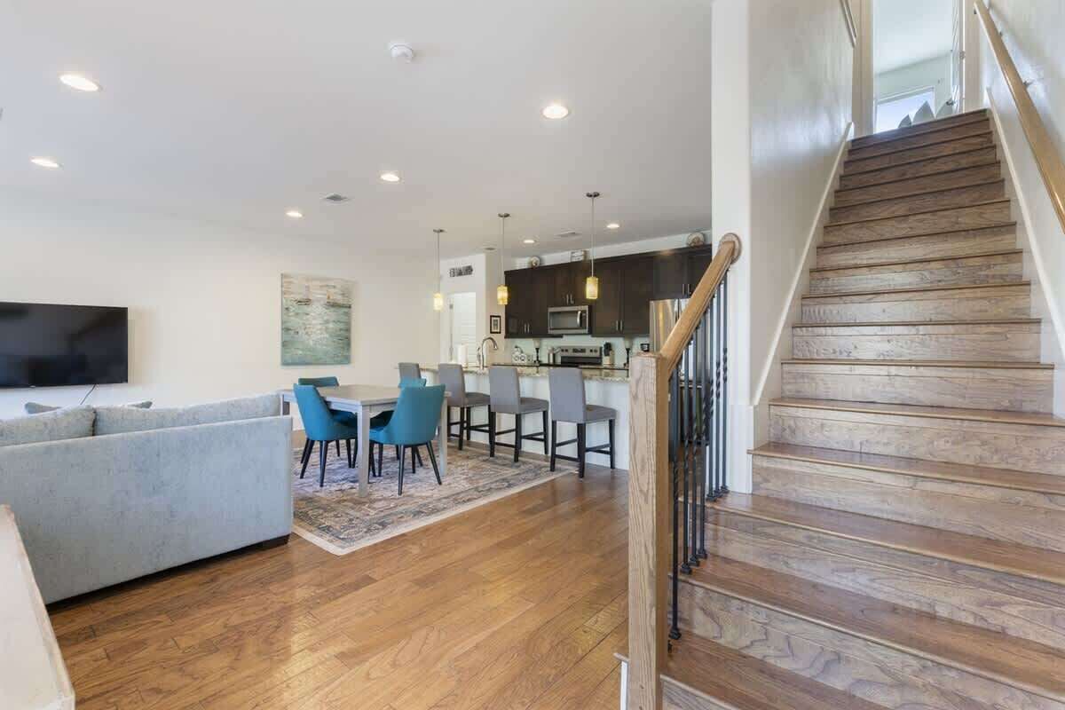 A modern living room with a sofa, dining table, and chairs, a kitchen area, and a wooden staircase.