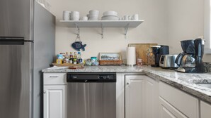 A kitchen with a stainless steel refrigerator, white cabinets, a dishwasher, a coffee maker, and a shelf with dishes and a framed picture.