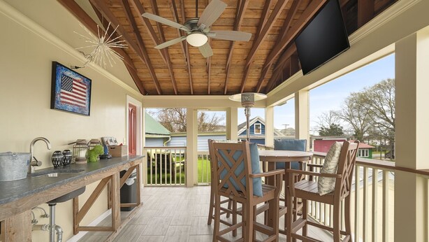 A covered patio area with a wooden ceiling, a ceiling fan, a TV mounted on the wall, and a view of neighboring houses.