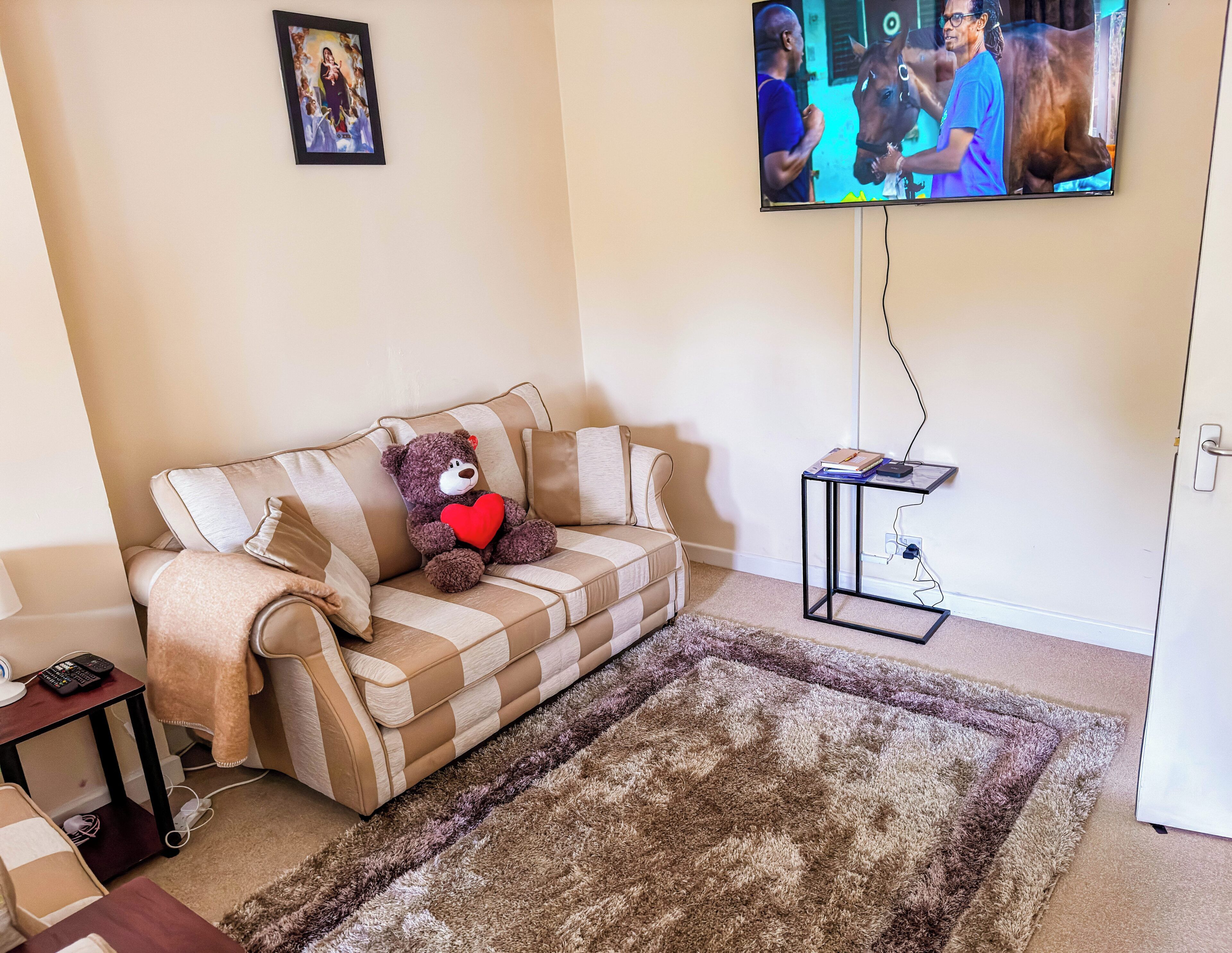 A living room with a beige sofa, a brown teddy bear with a red heart, a television mounted on the wall, and a small side table with a remote control.