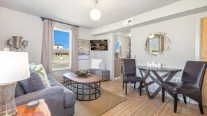 Una sala de estar moderna con un sofá gris, una mesa de centro de madera, una mesa de comedor con sillas y un ventanal con cortinas.