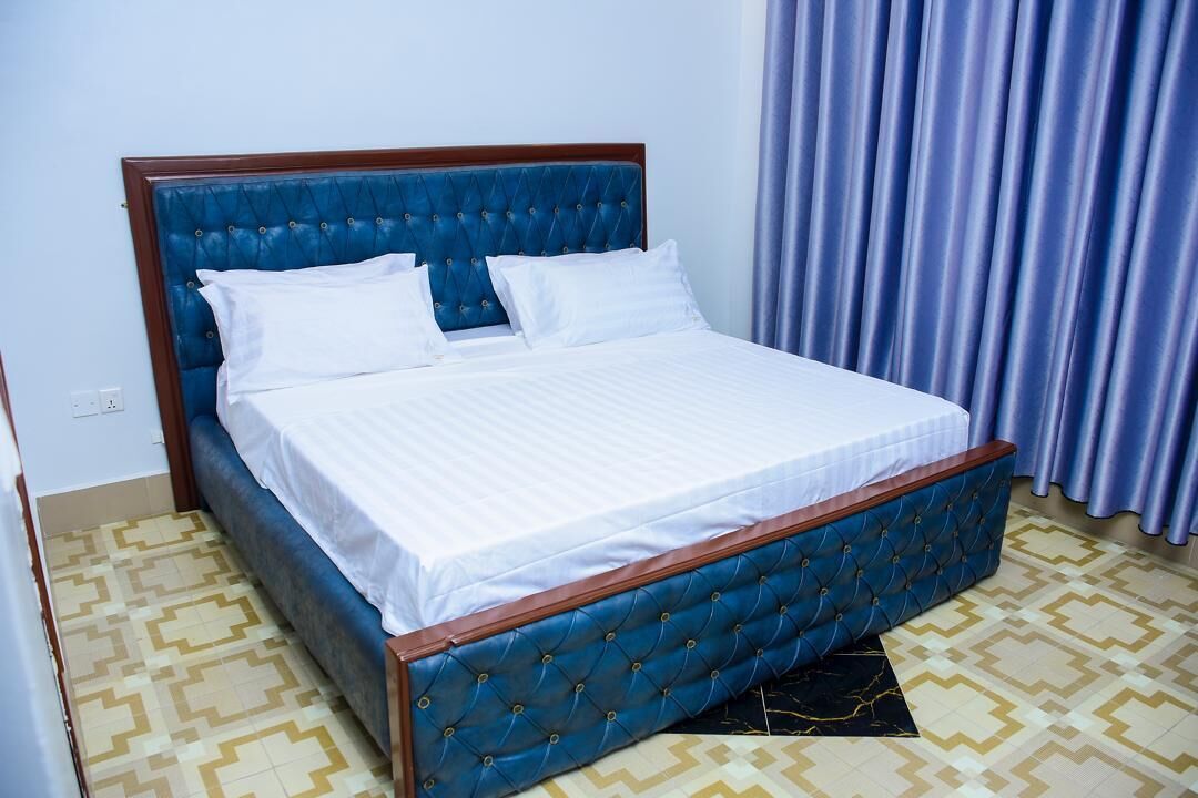 Ein Bett mit blauem gepolstertem Kopfteil und weißer Bettwäsche in einem Zimmer mit gemustertem Boden und einem Vorhang.