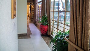 Pasillo de hotel con ventanas amplias, una maceta roja decorativa y plantas en macetas.