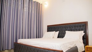 Una cama bien hecha con cabecera capitonada, una mesita de noche de madera y un gran ventanal con cortinas.