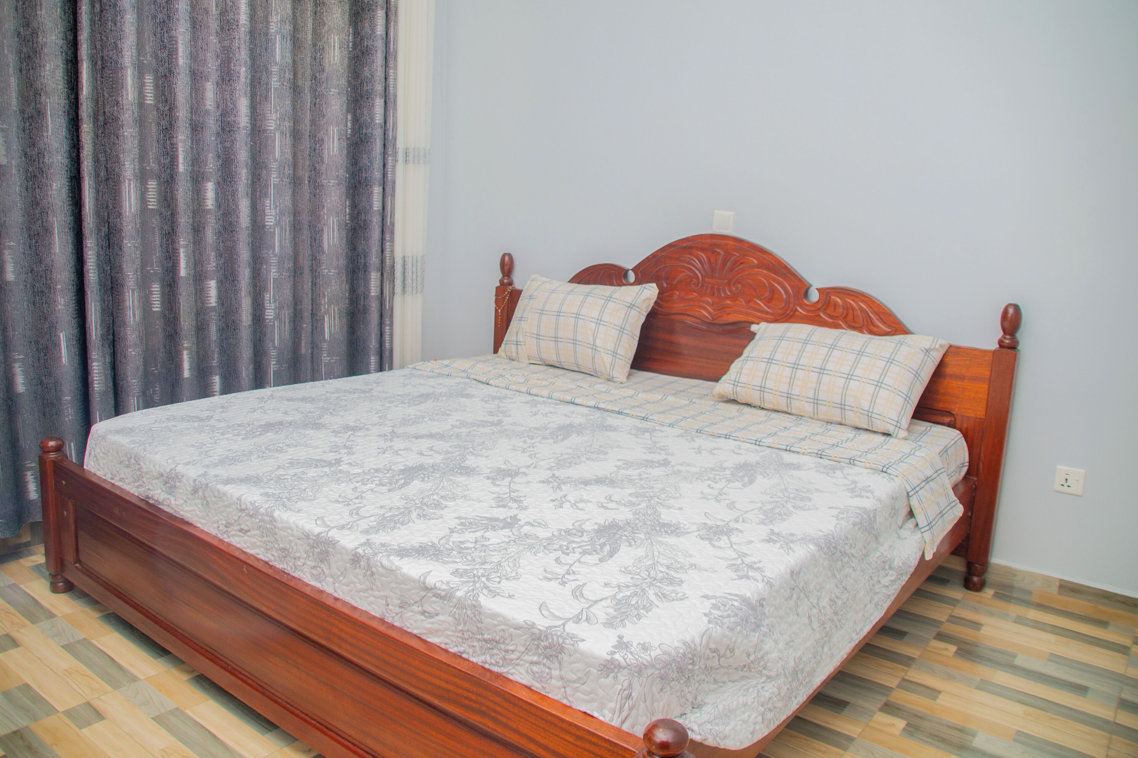 Una cama de madera con una colcha estampada con motivos florales y dos almohadas, piso de madera y una ventana con cortinas.
