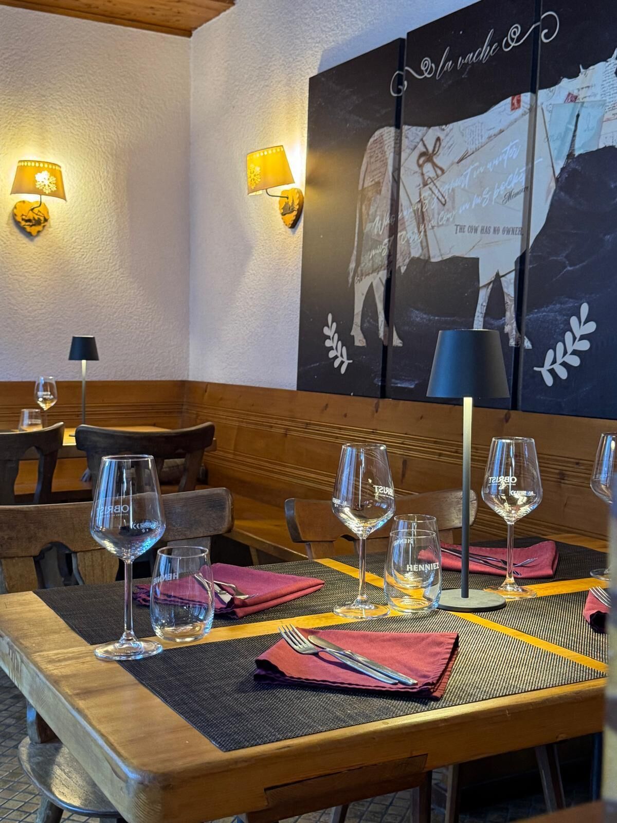 L’intérieur d’un restaurant avec des tables en bois, des verres à vin en verre transparent et un mur orné d’une affiche représentant une vache et un texte.
