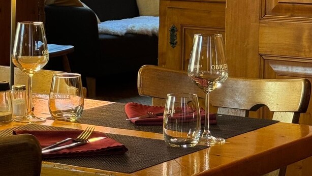 A dining area with a wooden table set for two, a window with curtains, and a chalkboard with writing. - Hotel La Tour d'Aï (Leysin)
