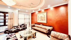 A living room with a glass coffee table, two sofas, a patterned rug, a chandelier, and a large framed picture on the wall. - South Inn – Green Park South Delhi (New Delhi)