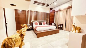 A modern bedroom with a large bed, a wooden headboard, a ceiling fan, and a small table with a chair. - South Inn – Green Park South Delhi (New Delhi)
