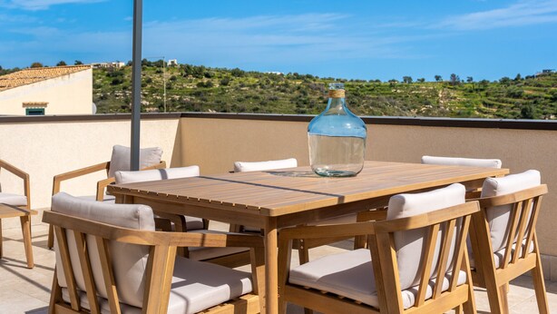 A wooden table with white cushioned chairs and a clear glass bottle on it, set on a balcony with a view of a hilly landscape. - Casa Verga con Terrazza by Wonderful Italy (Noto)