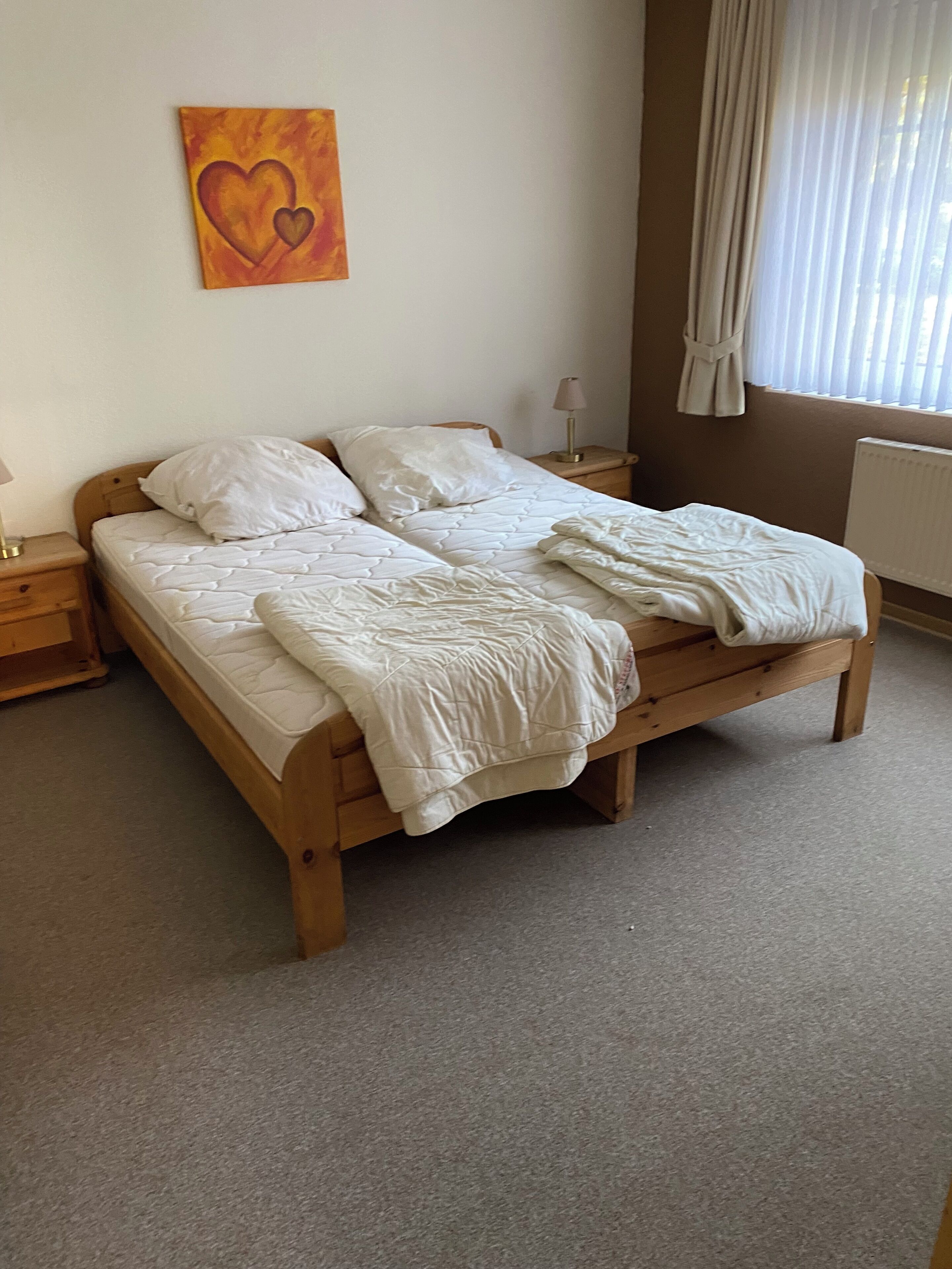 A bedroom with a wooden bed, white bedding, a nightstand with a lamp, and a painting of a heart on the wall.