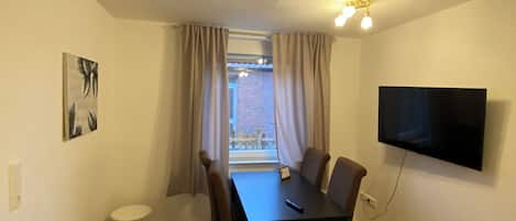 A room with a dining table, chairs, a small stool, a TV mounted on the wall, and a window with curtains.