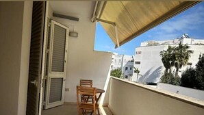 A balcony with a wooden chair and table, a sliding door, and a view of neighboring buildings and greenery. - Room-andromeda Rooms - Fenice (Otranto)