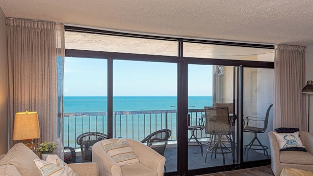 Une pièce dotée d’une grande porte-fenêtre coulissante donnant sur un balcon avec vue sur la mer. On y trouve deux canapés et une petite table avec une lampe.