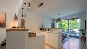 A modern kitchen with white cabinetry, a wooden countertop, and pendant lighting. There is a dining area with a table and chairs, and a large window offering a view of greenery outside. - Apartment in Duinbergen Near the Beach (Knokke-Heist)