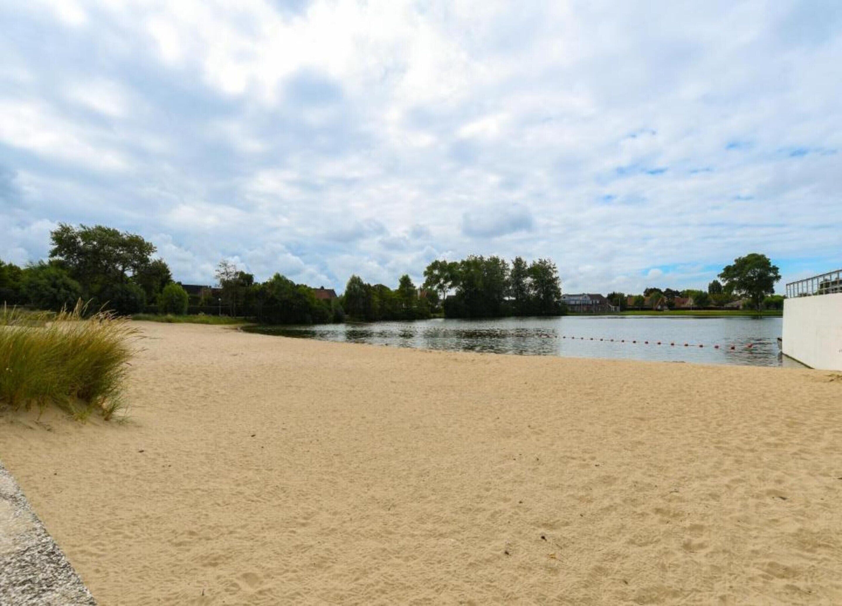 A sandy beach area with a calm water body, surrounded by trees and a cloudy sky.