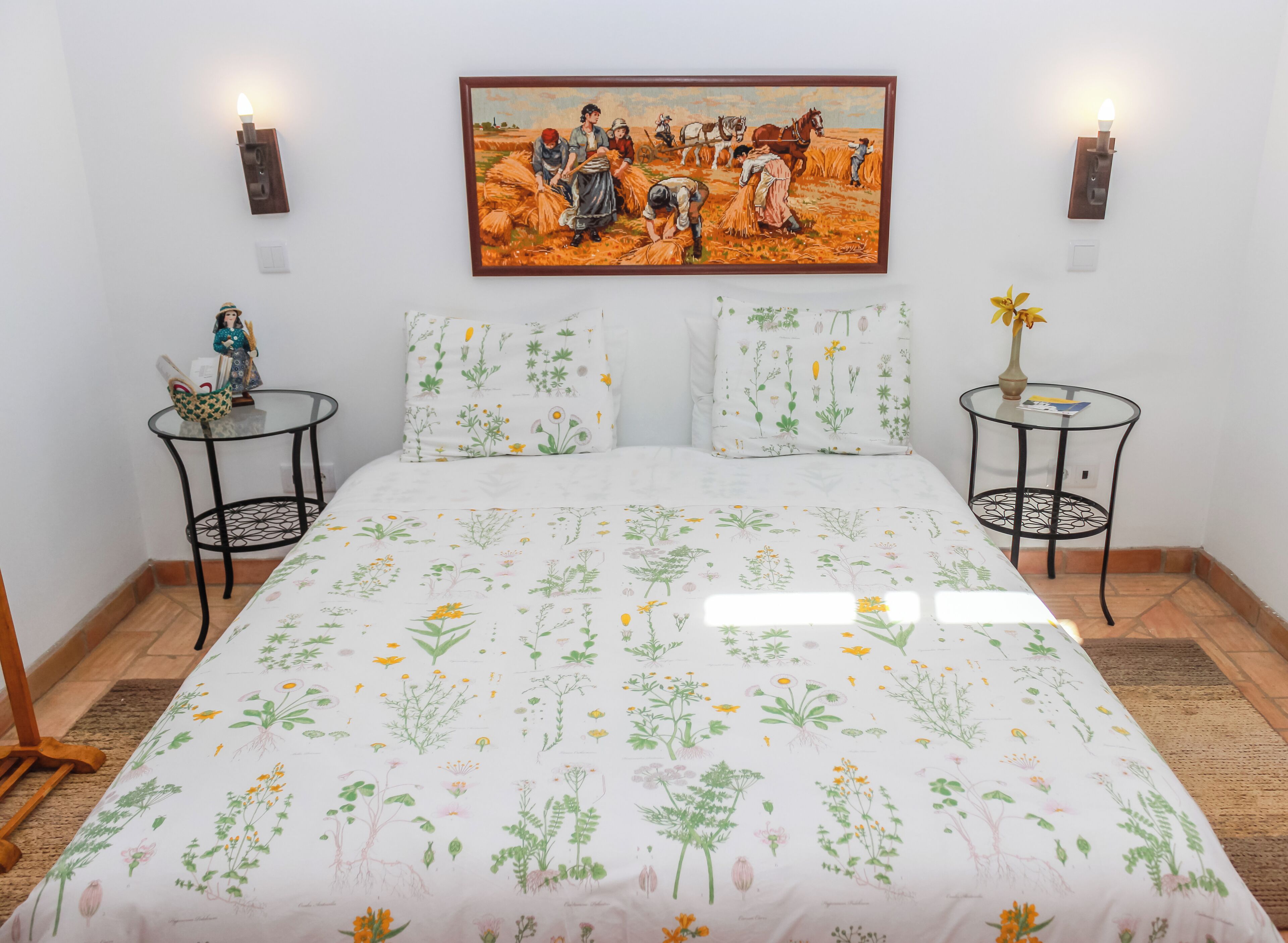 Un dormitorio con colcha estampada, dos mesitas de noche con adornos, un cuadro en la pared y una pequeña mesa redonda con un florero.