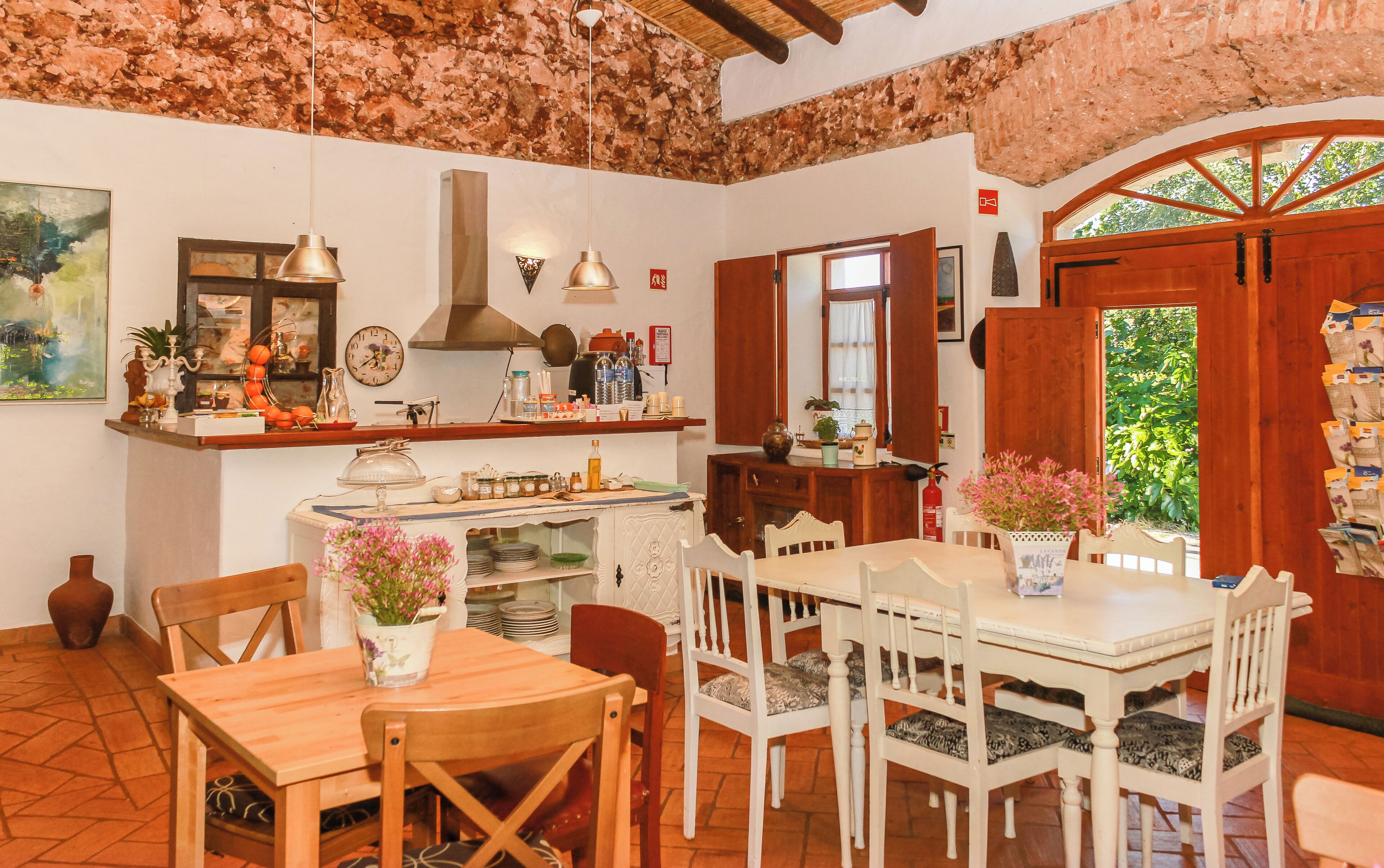 Zona d'esmorzar rústica amb taula i cadires de fusta, una barra de cuina amb diversos objectes i vista a la vegetació a través d'una porta de fusta.