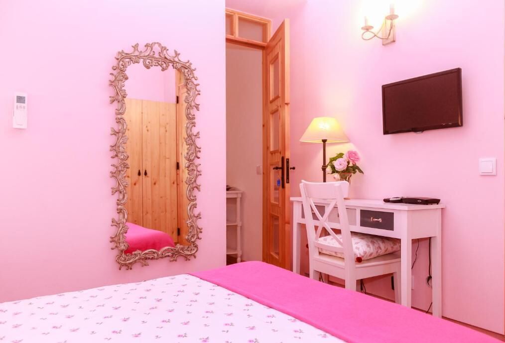 A bedroom with a bed, a desk with a chair, a mirror, a TV, and a lamp.