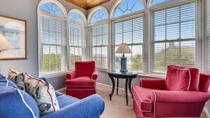 Un solarium con techos altos, ventanas arqueadas grandes, un sofá azul, un sillón rojo y una pequeña mesa con una lámpara.