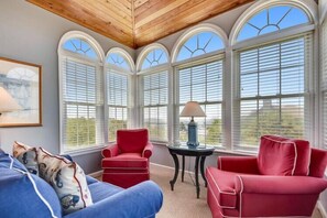 Una veranda con soffitto alto, ampie finestre ad arco, un divano blu, una poltrona rossa e un tavolino con una lampada.