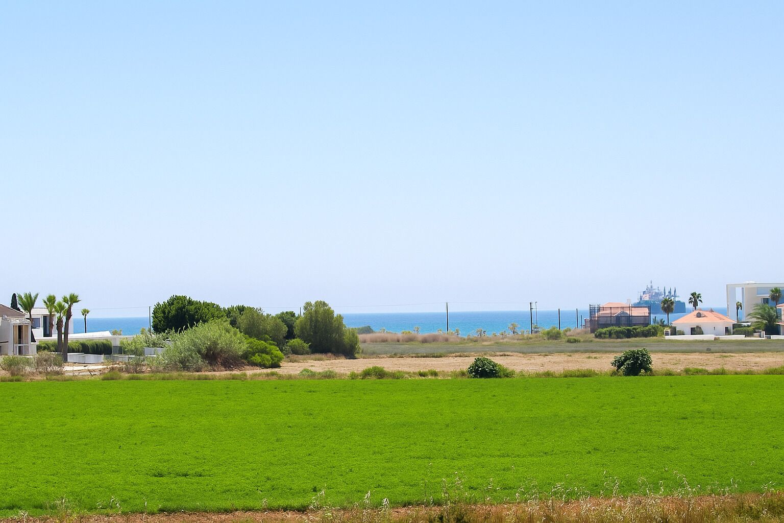 Un paysage côtier avec un champ herbeux, quelques bâtiments et une vue dégagée sur la mer.