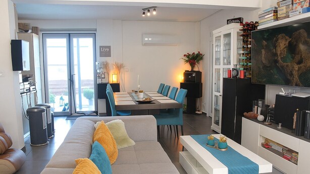 Een moderne woonkamer met een bank, eettafel en stoelen. Er is een keuken met kasten en een televisie.