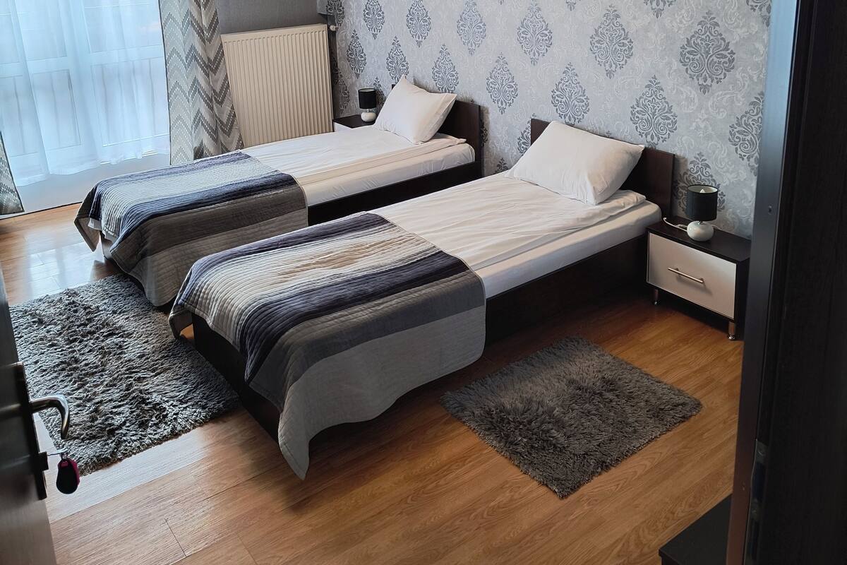 A bedroom with two beds, patterned wallpaper, a radiator, a nightstand, and a lamp.