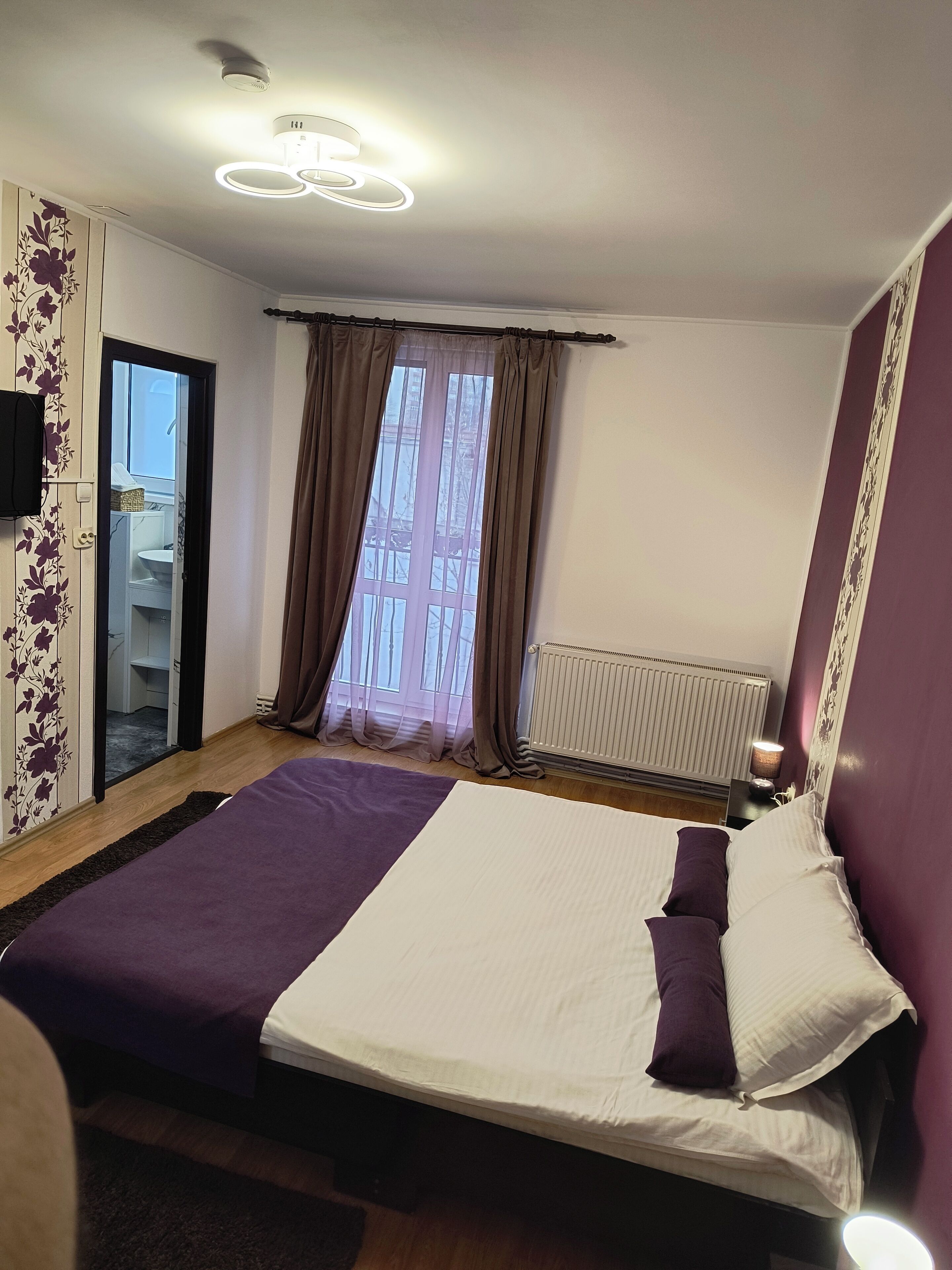 A bedroom with a bed, a window with curtains, a radiator, and a wall-mounted light fixture.