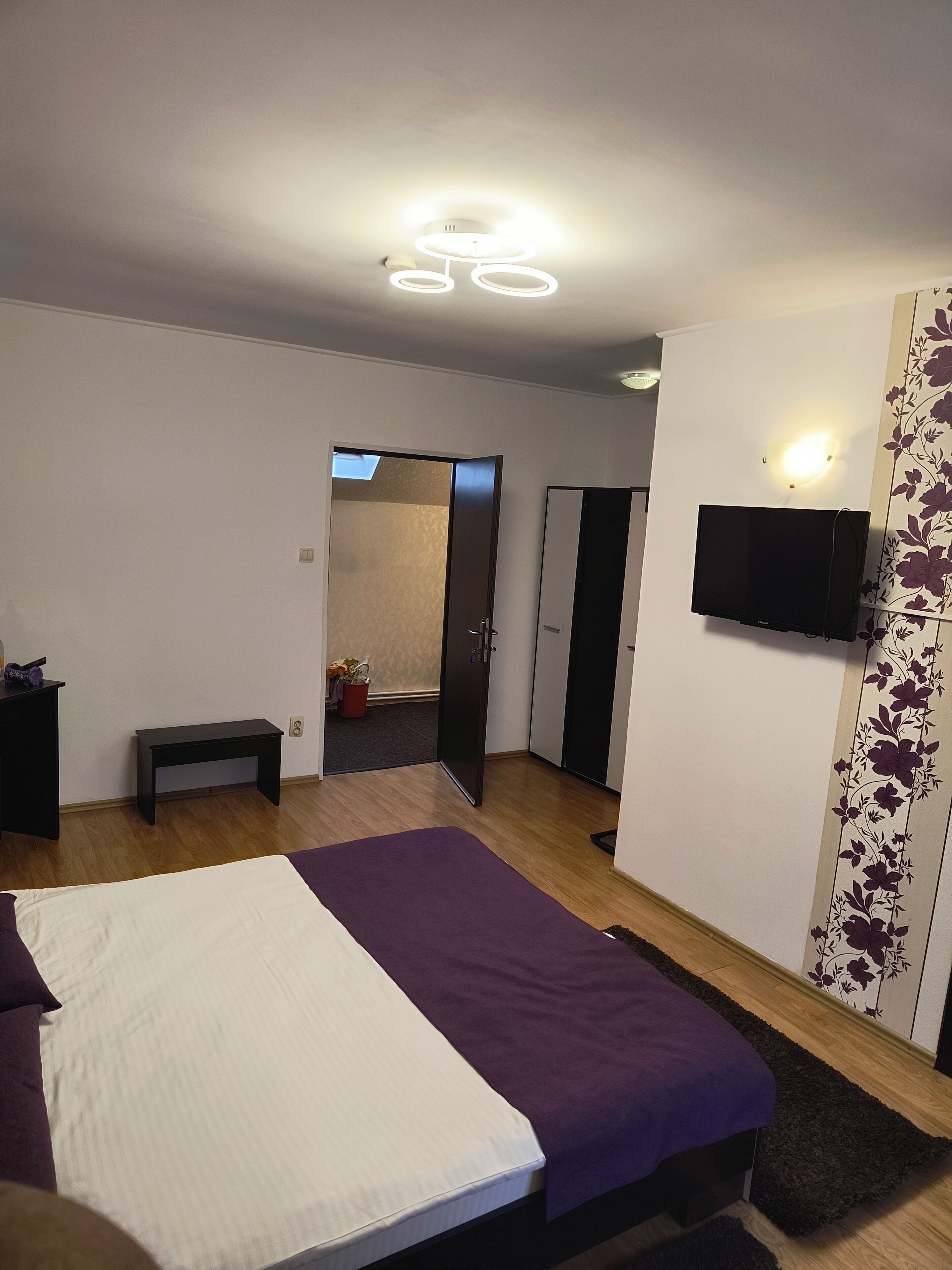 Un dormitorio con una cama, un banco, un televisor montado en la pared y una puerta que da a otra habitación.