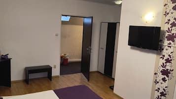 Un dormitorio con una cama, un banco, un televisor montado en la pared y una puerta que da a otra habitación.