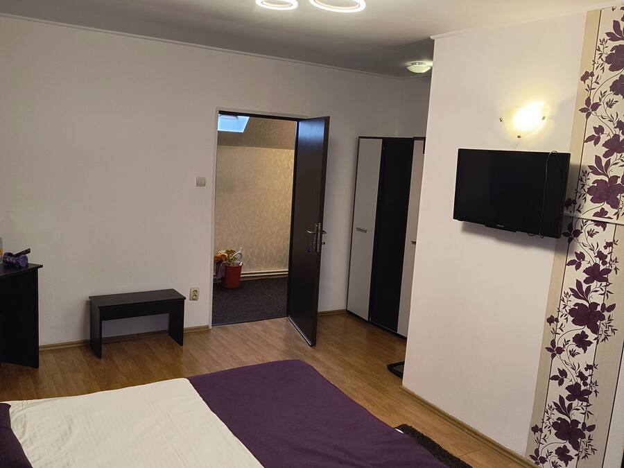 Un dormitorio con una cama, un banco, un televisor montado en la pared y una puerta que da a otra habitación.