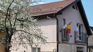 Un edificio de dos pisos con techo de tejas rojas, un balcón con una bandera y una entrada protegida por una reja.