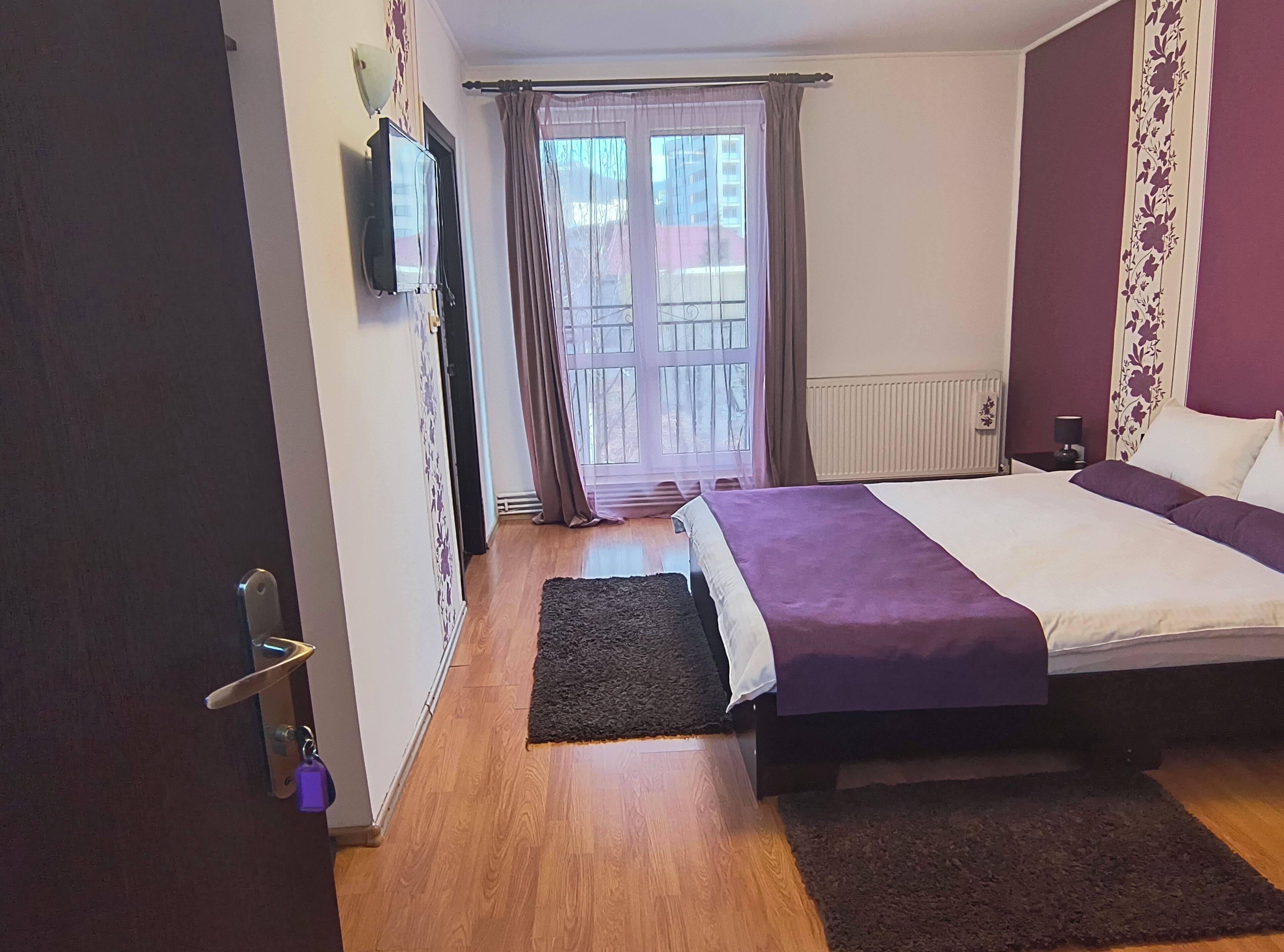 A bedroom with a bed, a TV mounted on the wall, a window with curtains, and a radiator.