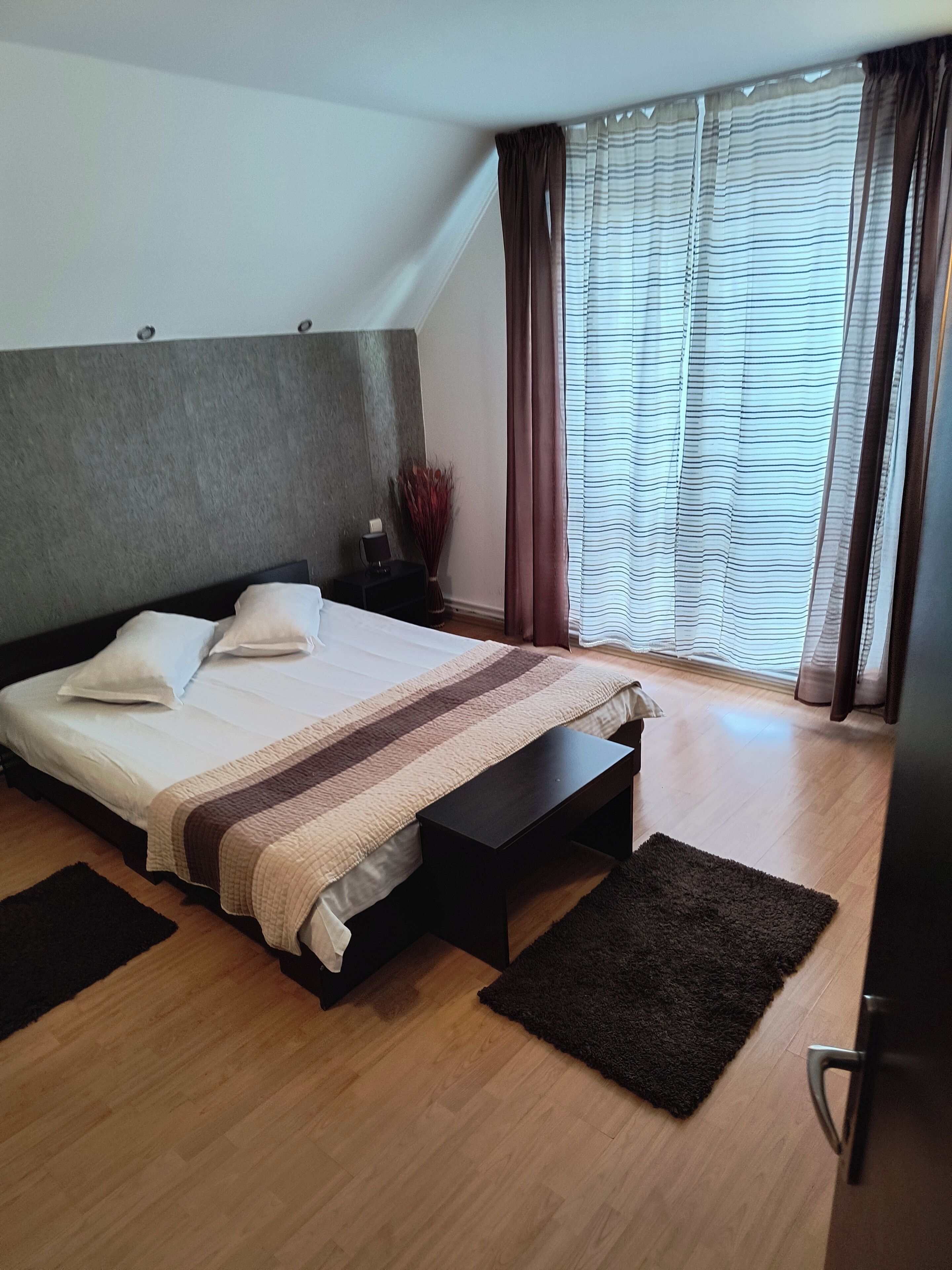 A modern bedroom with a large bed, a nightstand, a bedside table, a dark rug, and a window with curtains.