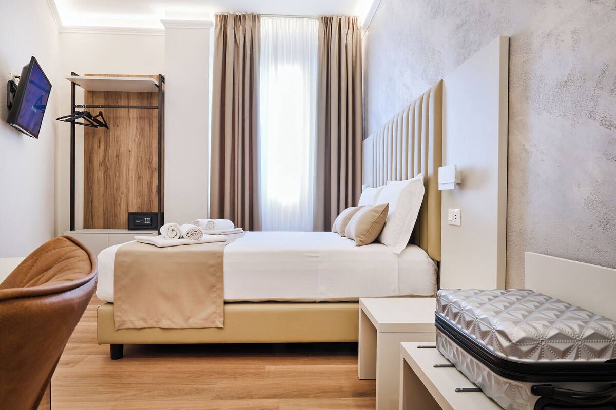 A modern hotel room with a bed, a desk, a chair, a wardrobe, and a suitcase.