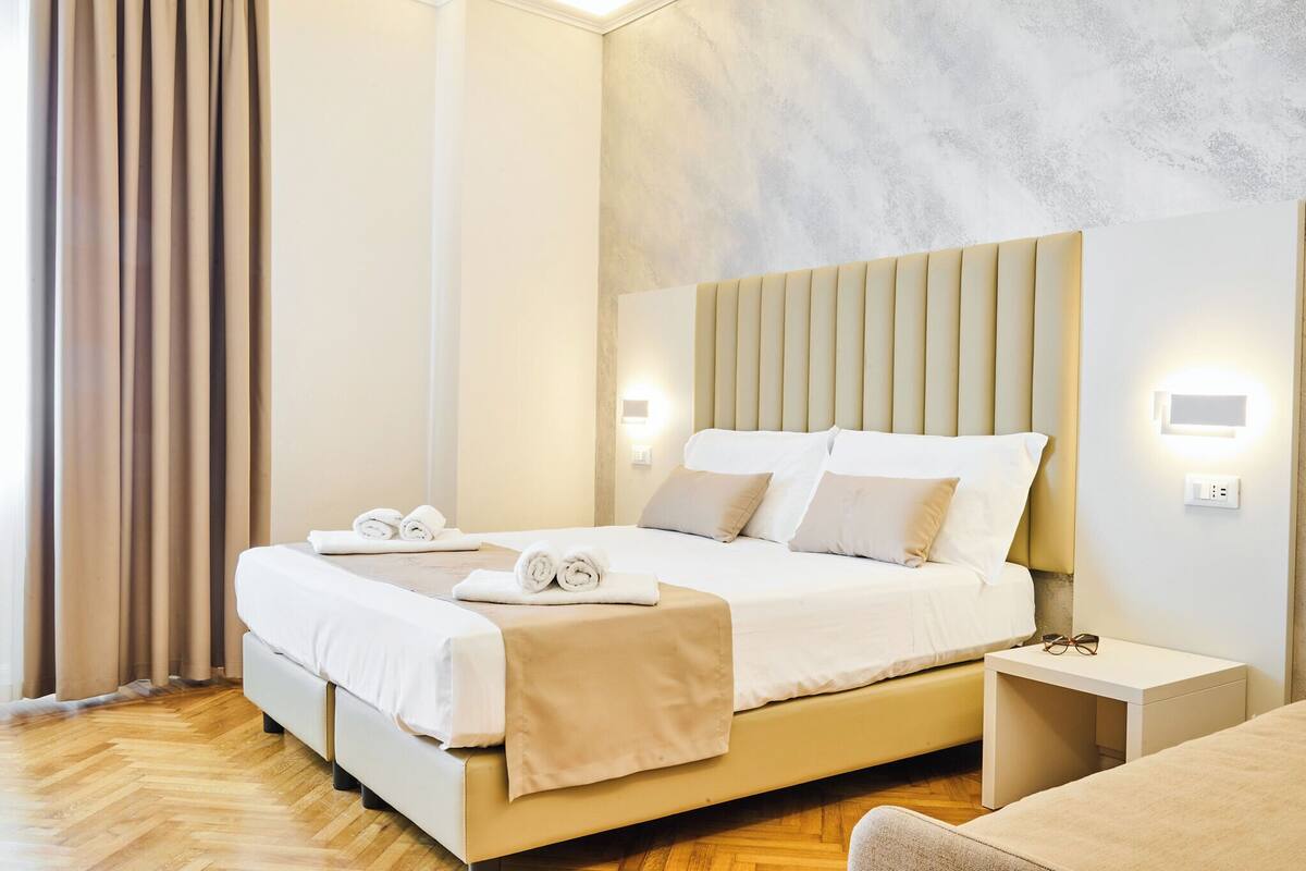 A neatly made bed with white linens and a beige throw blanket, a bedside table with glasses, and a wall-mounted light fixture.