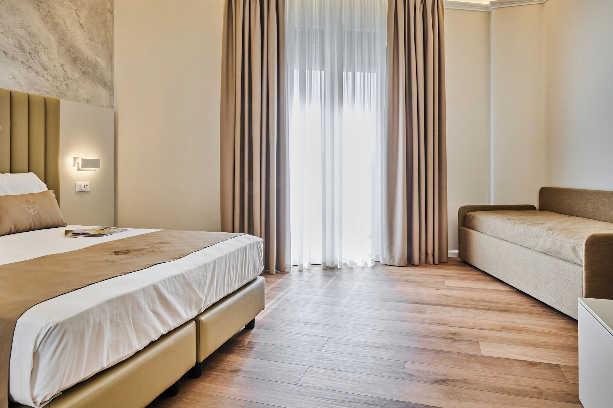 A modern hotel room with a large bed, a sofa, wooden flooring, and sheer curtains.