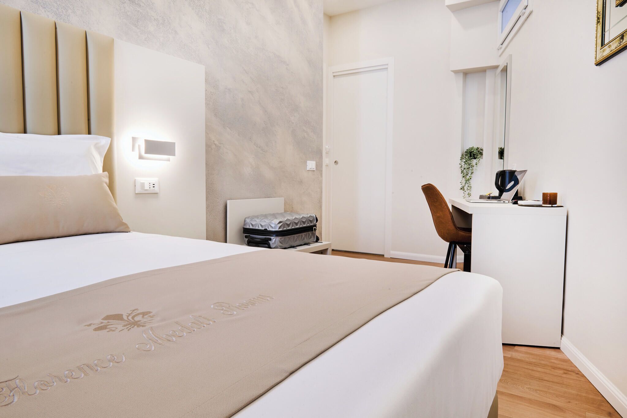 A hotel room with a bed, a desk, a chair, a suitcase, and a small plant.