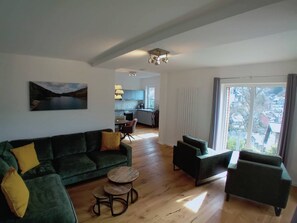 Een ruime zithoek met een groene bank, een ronde houten salontafel en uitzicht op een keuken en stadsbeeld door een groot raam.