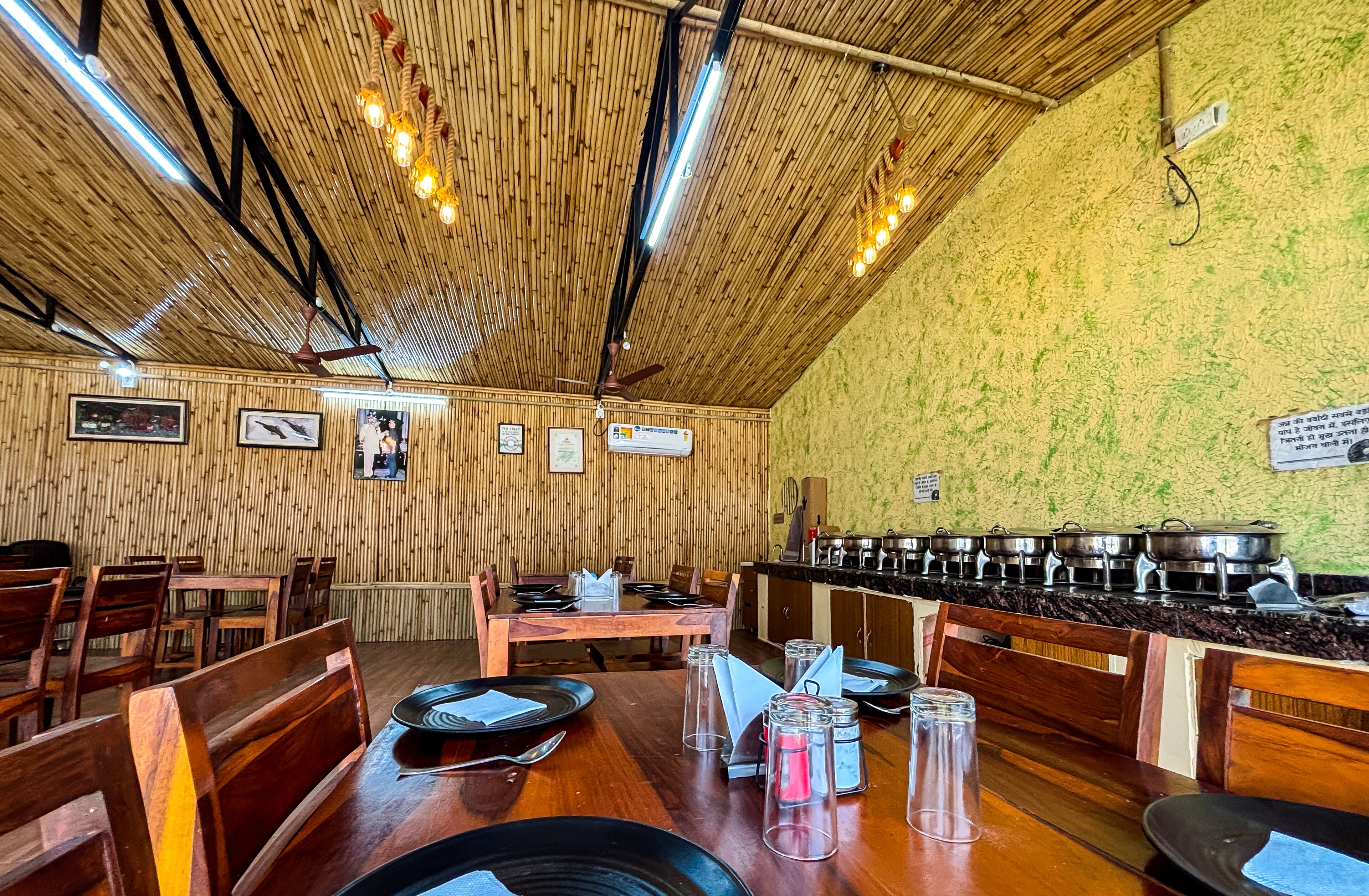 A restaurant with bamboo ceiling, wooden tables, and chairs. There are several framed pictures on the wall and a kitchen area with pots and pans.