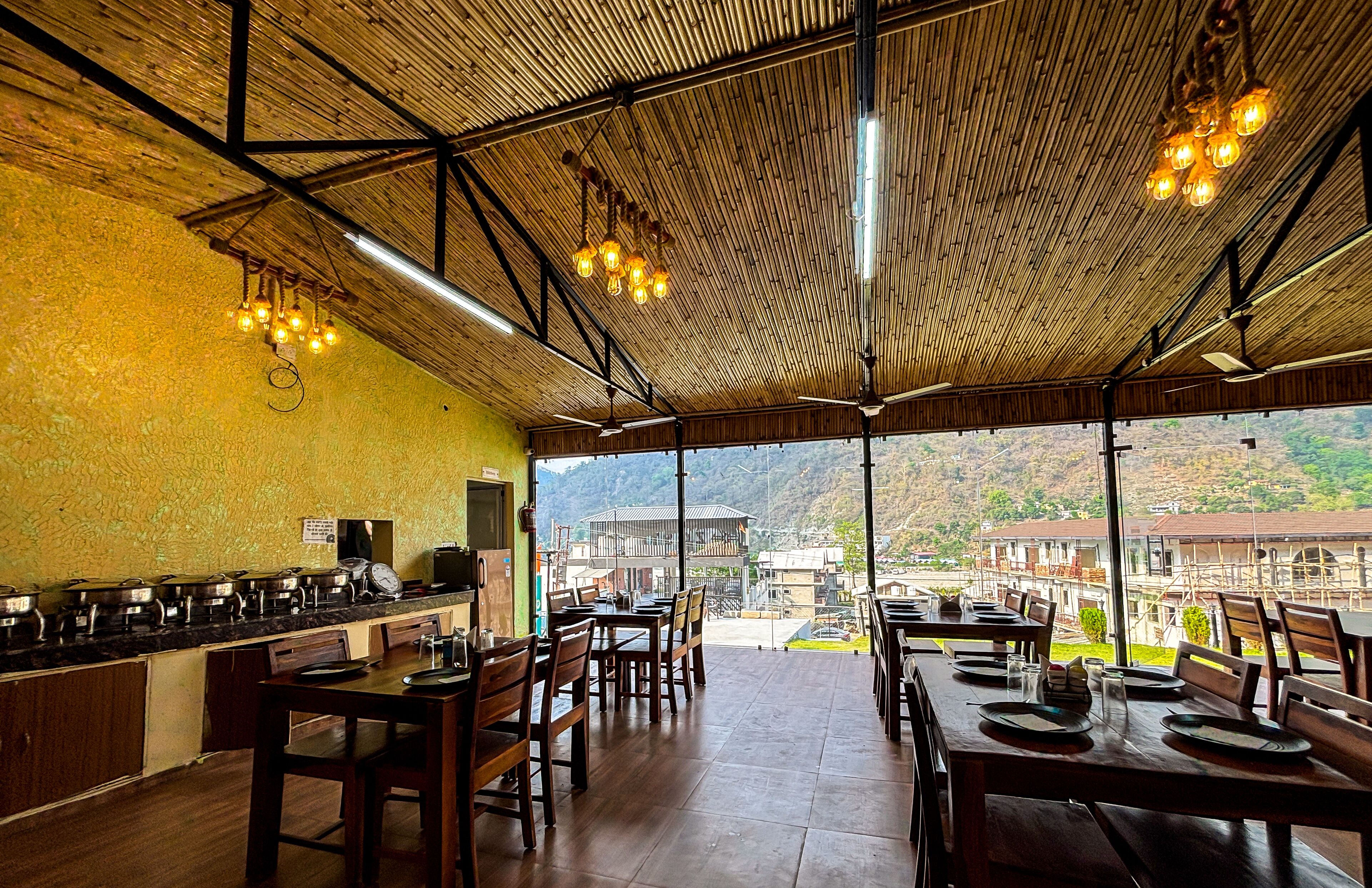 A restaurant with a bamboo ceiling, wooden tables and chairs, and a view of buildings and hills in the background.